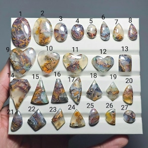 May include: A collection of twenty-seven polished cabochon gemstones arranged on a white display tray. Each stone features unique inclusions with shades of purple, orange, brown, and white. The stones are numbered 1 through 27 in black text.