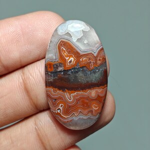 May include: Oval-shaped gemstone with a layered pattern of red, brown, gray, and white. The polished stone is held in a hand, showcasing the natural colors and unique veining. The gemstone is likely used for jewelry or decorative purposes.