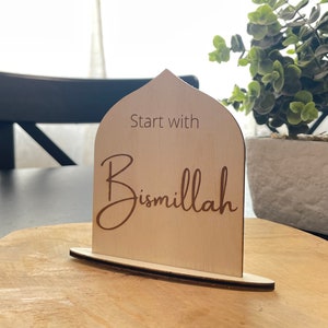 Op de afbeelding: Een lichtgekleurd houten bord met een gewelfde bovenkant en een voet. Het bord bevat de woorden "Start with Bismillah" in een scriptlettertype. Het bord staat op een houten oppervlak.