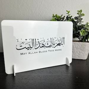 Op de afbeelding: Een wit acryl bord met zwarte Arabische kalligrafie en de tekst "Moge Allah dit huis zegenen".
