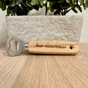 May include: A wooden bottle opener with a metal cap opener. The handle is engraved with the text "Der Austrincker".