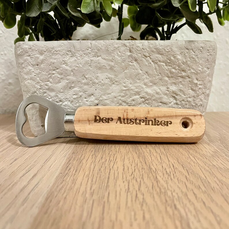 May include: A wooden bottle opener with a metal cap opener. The handle is engraved with the text "Der Austrincker".