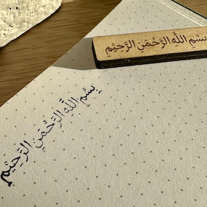 Könnte beinhalten: Ein weißes Notizbuch mit schwarzen gepunkteten Linien und dem arabischen Satz "بسم الله الرحمن الرحيم" in schwarzer Tinte geschrieben. Ein Holz-Lesezeichen mit dem gleichen Satz liegt auf dem Notizbuch.