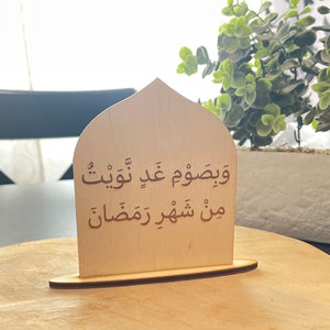 Op de afbeelding: Een houten bord in de vorm van een boog met de Arabische tekst "ويصوم عن طيب" en "من شهور رمضان" in bruine letters.