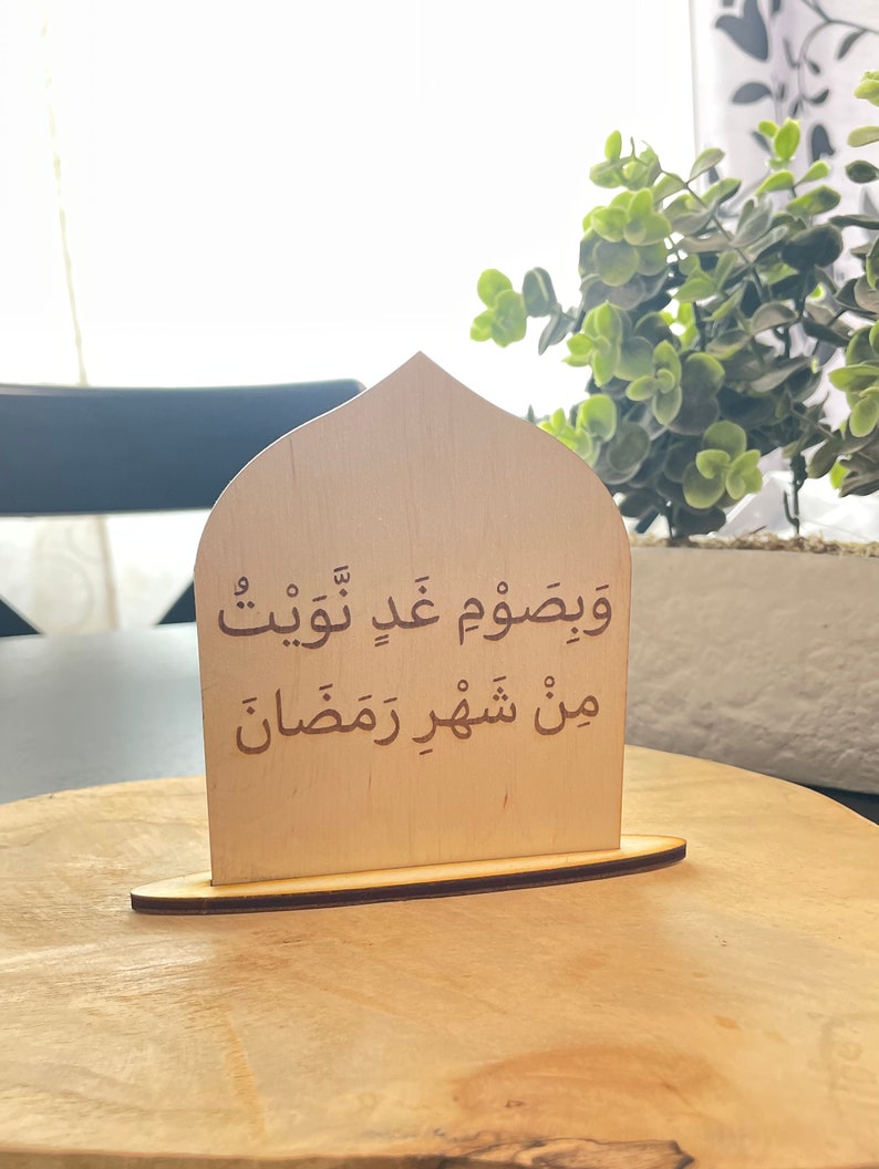 Op de afbeelding: Een houten bord in de vorm van een boog met de Arabische tekst "ويصوم عن طيب" en "من شهور رمضان" in bruine letters.