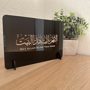 Op de afbeelding: Zwarte acrylplaat met gouden Arabische kalligrafie en de tekst "Moge Allah dit huis zegenen".