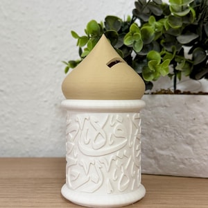Può includere: Un salvadanaio in ceramica bianca con calligrafia araba, sormontato da un coperchio a forma di cono color beige. La fessura per le monete è visibile sul coperchio. Il salvadanaio è su una superficie di legno, con una pianta verde sullo sfondo.