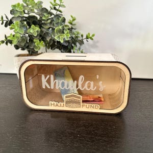 Può includere: Una scatola di legno con una finestra di plastica trasparente, etichettata "Hajj Fund" e personalizzata con il nome "Khawla's". La scatola è progettata per contenere denaro per un pellegrinaggio alla Mecca.