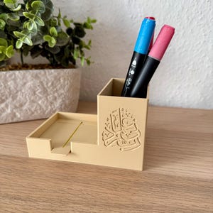 Könnte beinhalten: Ein beigefarbener Schreibtisch-Organizer mit Stifthalter und kleinem Fach. Der Stifthalter enthält einen blauen und einen rosa Marker. Der Organizer hat eine arabische Inschrift. Er steht auf einer Holzoberfläche.