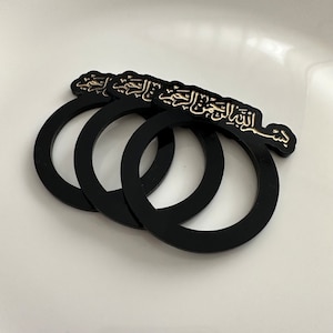 Könnte beinhalten: Set aus drei schwarzen Serviettenringen mit goldener arabischer Kalligraphie. Die Kalligraphie lautet "Im Namen Allahs, des Allerbarmers, des Barmherzigen".