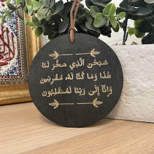 Op de afbeelding: Een ronde houten hanger met een zwarte achtergrond en gouden Arabische kalligrafie. De tekst luidt "Subhana alladhi sakhkhara lana hadha wa ma kunna lahu muqrinin wa inna ila rabbina lamunqalibun".