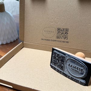 Puede incluir: Un sello de madera con un código QR y el texto "THIS PACKAGE IS HAPPY TO SEE YOU TOO!" se coloca en una caja de cartón. La caja está abierta, revelando el interior y el logotipo "BISMAA".