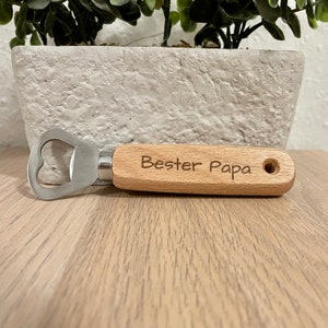 May include: A wooden bottle opener with a metal cap opener. The handle is engraved with the text "Bester Papa".