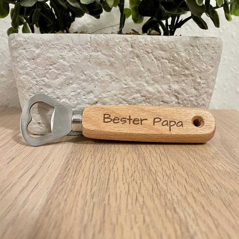 May include: A wooden bottle opener with a metal cap opener. The handle is engraved with the text "Bester Papa".