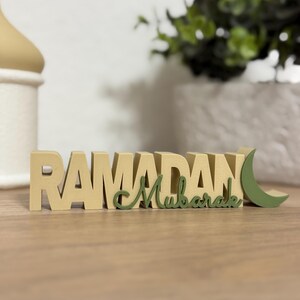 May include: A wooden Ramadan Mubarak sign in beige and green. The word "Ramadan" is in block letters, and "Mubarak" is in cursive. A green crescent moon is on the right side of the sign. The sign is on a wooden surface.