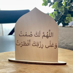 Op de afbeelding: Een houten bord met een gebogen bovenkant, met een islamitisch gebed in Arabisch schrift. De tekst luidt "Allahumma innaka afuwwun wa tuhibbul afwa".