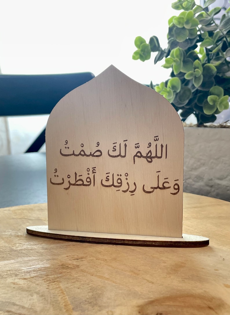 Op de afbeelding: Een houten bord met een gebogen bovenkant, met een islamitisch gebed in Arabisch schrift. De tekst luidt "Allahumma innaka afuwwun wa tuhibbul afwa".