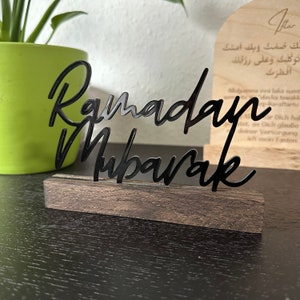 May include: Black wooden sign with the words "Ramadan Mubarak" in a stylised script. The sign is on a dark wood surface.