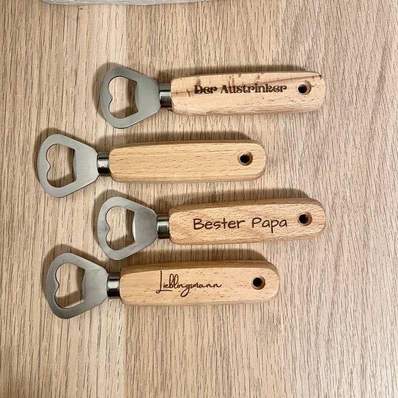 May include: Four wooden bottle openers with different engraved text. The text reads "Der Austrincker", "Bester Papa", "Lieblingsmann".