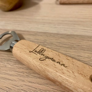 May include: A wooden bottle opener with a metal opener and a laser engraved inscription that reads "Lieblingsmann".