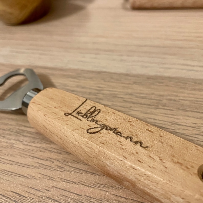 May include: A wooden bottle opener with a metal opener and a laser engraved inscription that reads "Lieblingsmann".