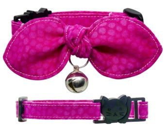 May include: A bright pink pet collar featuring a fabric bow with a subtle tonal dot pattern and white stitching. A silver-toned metal bell hangs from the center of the bow. The collar includes a black plastic buckle shaped like a cat face.