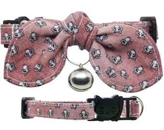 May include: A pink cat collar with a bow and a bell. The bow has a floral pattern and the collar has a black cat head buckle.