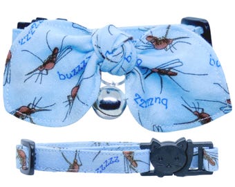 May include: A light blue pet collar featuring a bow tie with a repeating mosquito pattern and the text "buzzz". The collar includes a small silver bell attached to the center of the bow and a black plastic buckle shaped like a cat face.