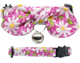 May include: A pink cat collar with a daisy floral pattern and a matching bow tie. The collar has a black breakaway buckle with a cat face design and a silver bell. The bow tie is attached to the collar.