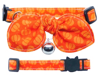 May include: An orange pet collar featuring a repeating pumpkin pattern, a large fabric bow tied at the center, a small silver metal bell, and black plastic buckles. The collar includes an adjustable strap with white stitching along the edges.