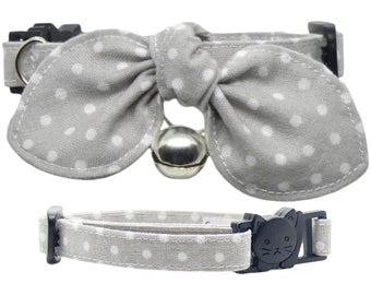 May include: A gray cat collar with white polka dots and a matching bow tie. The collar has a silver bell and a black buckle with a cat face design. The collar is designed for cats.