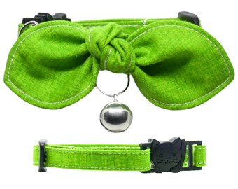 May include: A green cat collar with a bow and a bell. The collar has a black buckle with a cat face on it.