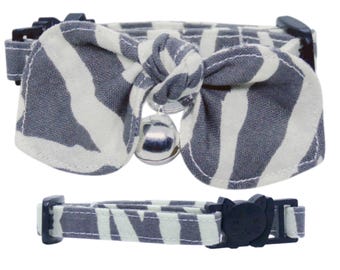 May include: A pet collar featuring a grey and white zebra-striped fabric bow and a small silver-toned metal bell. The collar includes a dark navy blue plastic buckle shaped like a cat head and an adjustable strap with a matching zebra-striped pattern.