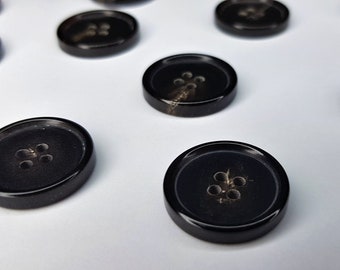 Könnte beinhalten: Ein Satz schwarzer Knöpfe mit jeweils vier Löchern. Die Knöpfe haben eine leicht glänzende Oberfläche und sind aus einem glatten Material gefertigt.