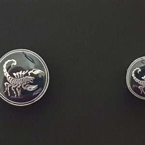 May include: Two silver-toned buttons with a black background and a white scorpion design. The larger button is slightly larger than the smaller button.