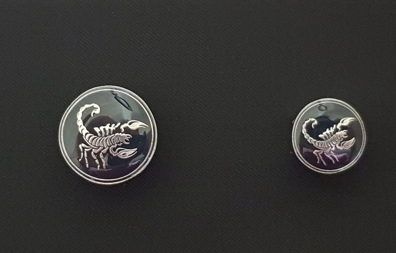 May include: Two silver-toned buttons with a black background and a white scorpion design. The larger button is slightly larger than the smaller button.
