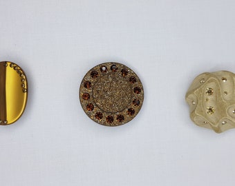 May include: Three decorative buttons with gold and brown accents. The buttons are round and oval shaped with a sparkly finish.
