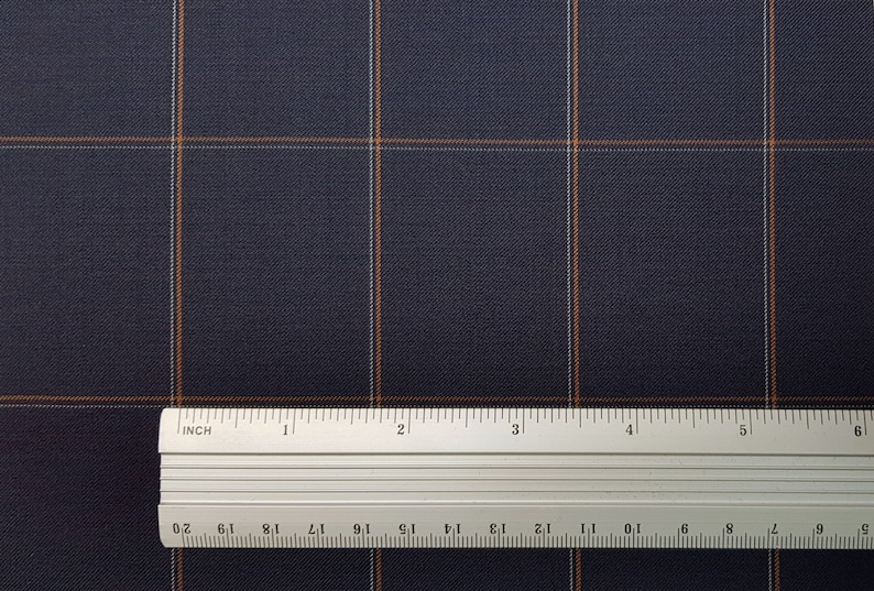 K&ouml;nnte beinhalten: Nahaufnahme eines dunkelblauen Stoffes mit einem dezenten orange-wei&szlig;en Karomuster. Ein Lineal liegt auf dem Stoff und zeigt die Skala in Zoll.