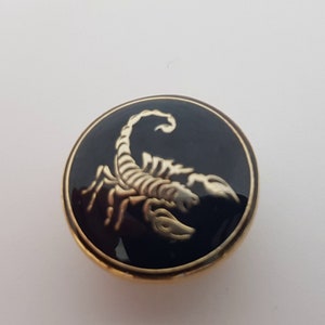 May include: A gold-colored button with a black enamel background featuring a gold scorpion design.