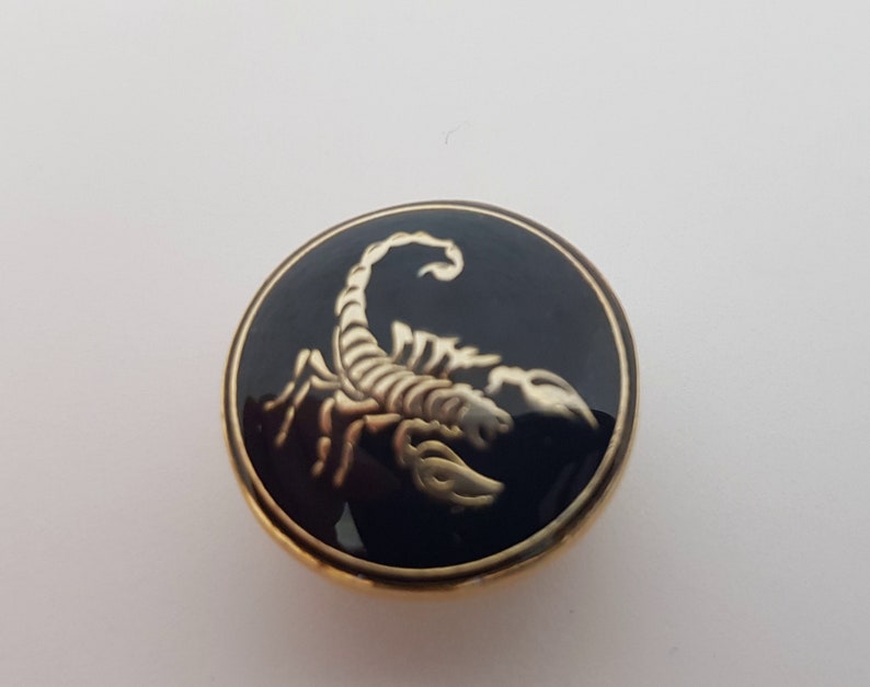 May include: A gold-colored button with a black enamel background featuring a gold scorpion design.