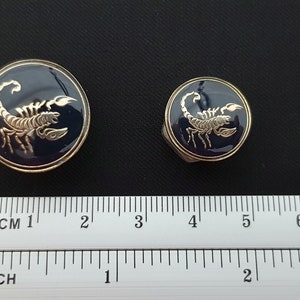 May include: Two gold-colored metal buttons with a blue enamel background and a gold scorpion design. The larger button is 1 inch in diameter and the smaller button is 3/4 inch in diameter.