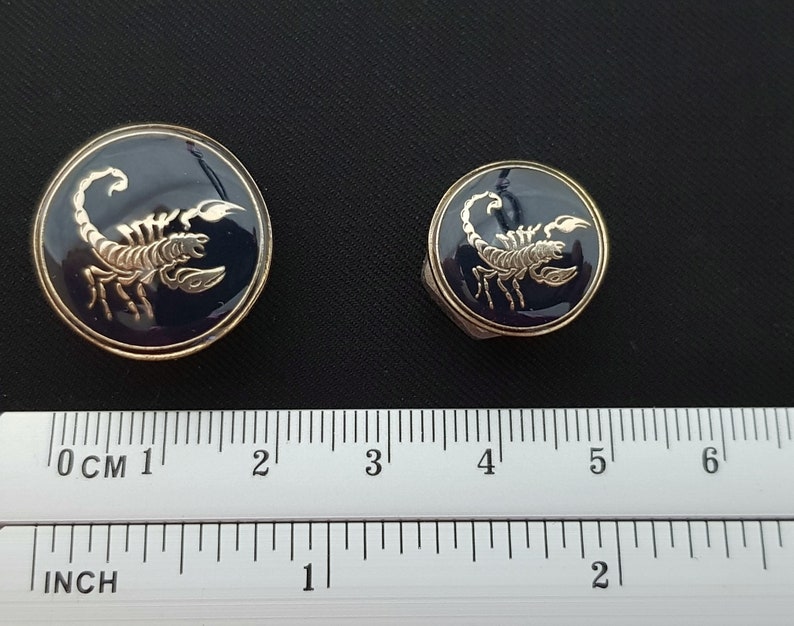 May include: Two gold-colored metal buttons with a blue enamel background and a gold scorpion design. The larger button is 1 inch in diameter and the smaller button is 3/4 inch in diameter.