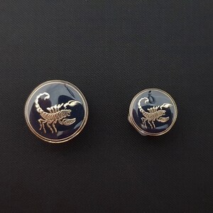 May include: Two round, blue, metal buttons with gold scorpions on them. The larger button is about 1 inch in diameter and the smaller button is about 1/2 inch in diameter.