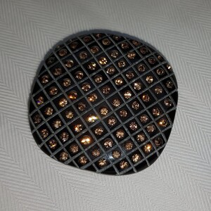 Puede incluir: Un broche de resina negra con un patrón de cuadrícula de pequeños diamantes de imitación marrones.