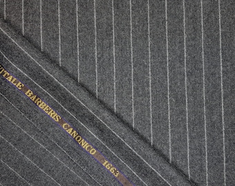 May include: A dark gray fabric featuring a white pinstripe pattern. A dark brown selvedge edge runs diagonally across the material, printed with the gold text VITALE BARBERIS CANONICO 1663. The textile has a visible weave and a folded corner.