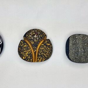 Puede incluir: Tres botones decorativos con diseños únicos. El primer botón es redondo y negro con un diseño de flor blanca. El segundo botón tiene forma de hoja con un diseño moteado dorado y negro. El tercer botón es cuadrado con un diseño moteado gris y negro.