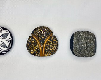 May include: Three decorative buttons with unique designs. The first button is round and black with a white flower design. The second button is a leaf shape with a gold and black speckled design. The third button is a square with a gray and black speckled design.
