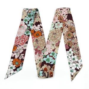 Könnte beinhalten: Ein langer, schmaler Schal mit dunkelbraunem Hintergrund und einem dichten, mehrfarbigen Blumenmuster. Das Design zeigt verschiedene Blumen in Orange-, Hellblau-, Rosa- und Weißtönen, durchsetzt mit kleinen Schmetterlingsillustrationen.