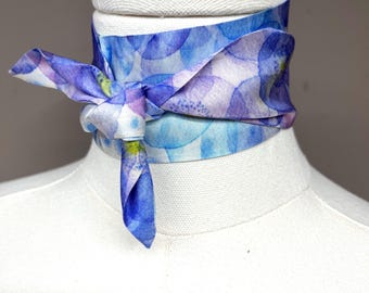 Puede incluir: Un pañuelo de seda con un estampado de acuarela azul y morado, atado alrededor del cuello de un maniquí. El pañuelo está anudado en la parte delantera, con los extremos colgando. El diseño presenta tonos de azul, morado y blanco.