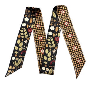 May include: A long, narrow scarf featuring a dual-pattern design. One section displays a black background with botanical illustrations of leaves, flowers, and berries in gold, white, and red. The other section features a black and gold checkered pattern.